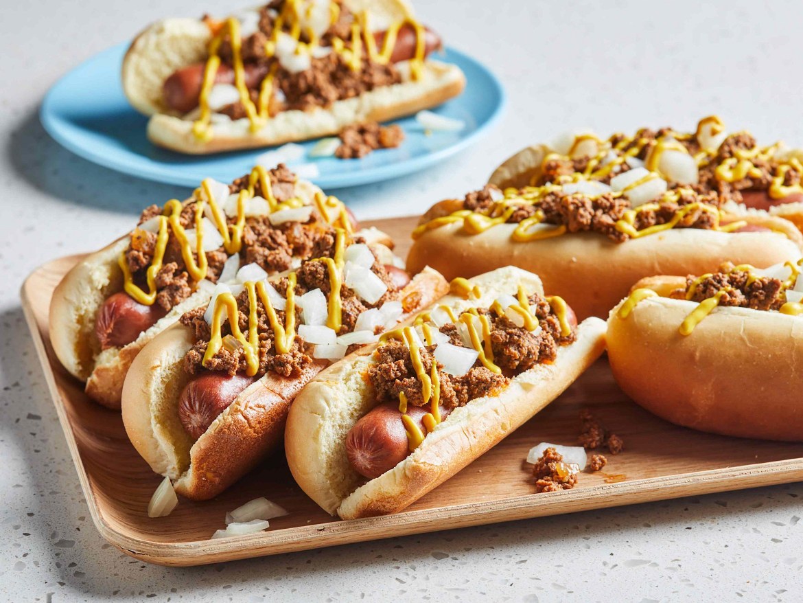 Beef hot dogs on a wooden board topped with chili, mustard and diced onions