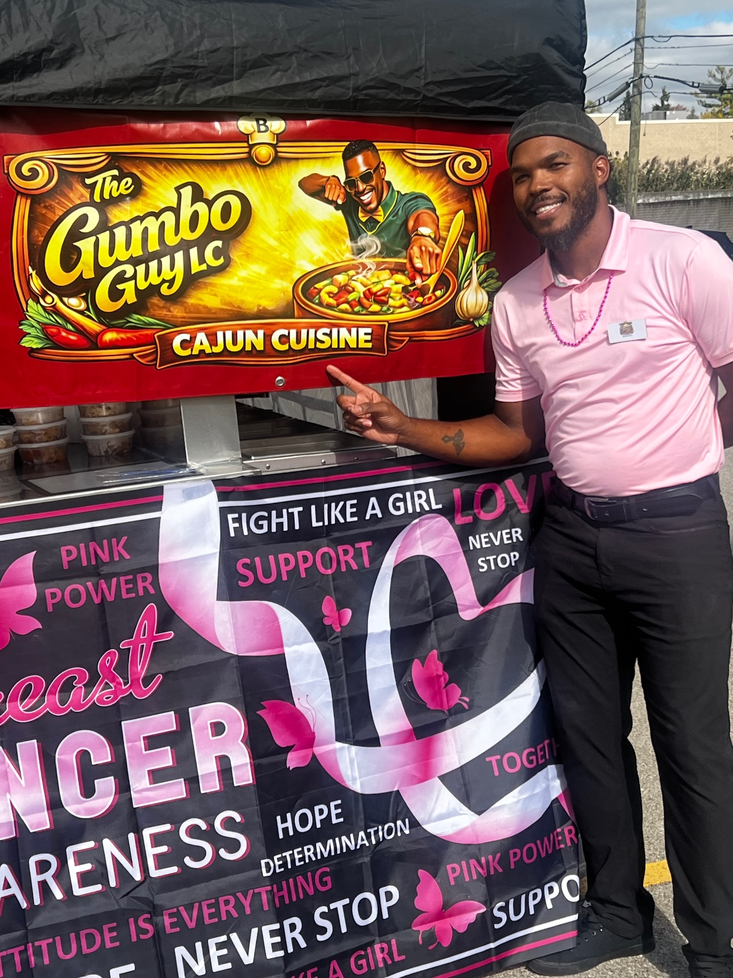 Brandel (The Gumbo Guy) pointing to The Gumbo Guy LC cart and its Breast Cancer Awareness banner